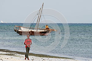 Teen boy walks on coast Indian Ocean, near fishing boat.