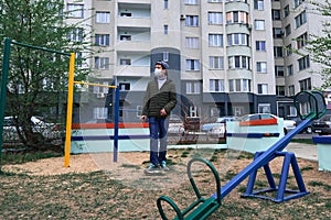 Teen boy walks through the playground near high-rise buildings with apartments, a residential area, a medical mask on his face