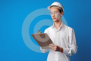 Teen boy in white hardhat writing on clipboard against blue background
