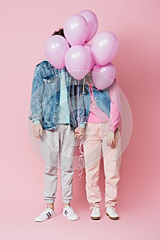 Teen couple holding hands while balloons covering faces on pink background.