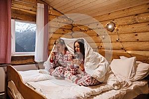 Teen friends in same Christmas pajamas relaxing in bed inside cozy log cabin with winter view