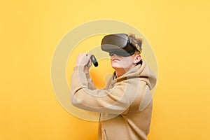 Teen gamer in a VR helmet stands on a yellow background with controls in his hands and plays virtual reality games, looking away