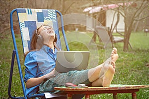 Teen girl barefoot laughing during online conference with her school friends sitting in a deckchair outdoors in springtime