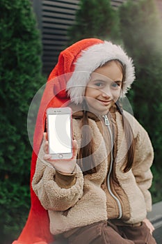 Teen girl in Christmas Santa hat shows phone with white screen. Chroma key white screen smartphone set up for advertising. Cute