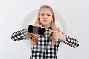 Teen girl with concerned face pointing at mobile phone, looks worried at camera, showing something upsetting on smartphone. Indoor