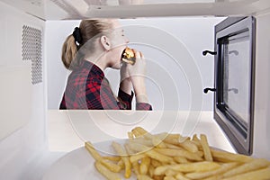 Teen girl greedily eats hamburger mouth wide open. Sitting at table near the microwave. View through open oven