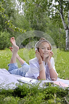 Teen girl lies listens to music in headphones on the grass in the park with books and notebooks