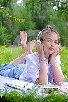 Teen girl lies listens to music in headphones on the grass in the park with books and notebooks