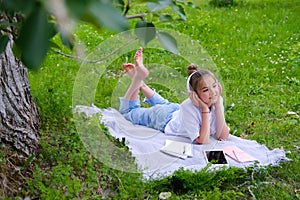 Teen girl lies listens to music in headphones on the grass in the park with books and notebooks