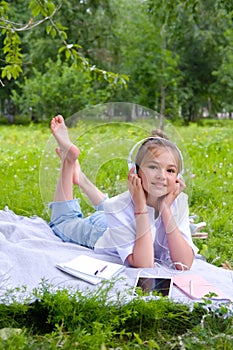 Teen girl lies listens to music in headphones on the grass in the park with books and notebooks