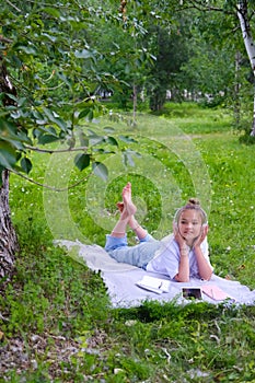 Teen girl lies listens to music in headphones on the grass in the park with books and notebooks