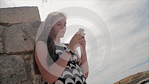 Teen girl listening to music with headphones on smartphone on a background of sea and the ruins of the ancient city