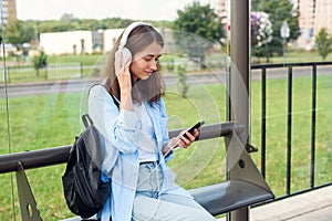 Teen girl listens to the music by white headphones in a public transport station while she waiting for tram.