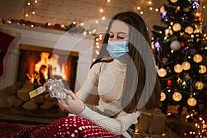 Teen girl in medical mask takes vitamins, sitting in front of decorated christmas tree and fireplace at home