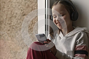 Teen girl with Mobile Phone and Headphones listening to music, audiobooks, audio lessons at window.  Online. Concept of Education