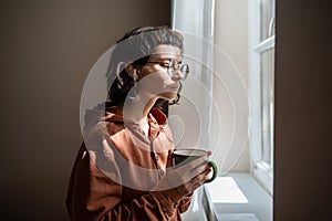 Teen girl nerd in glasses looking at window drinking cup of tea at home. Introvert, friendless