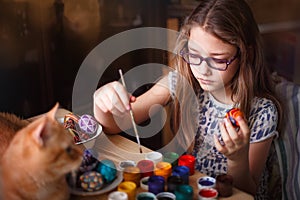 Teen girl paints Easter eggs, her ginger cat lies on the table