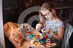 Teen girl paints Easter eggs, her ginger cat lies on the table