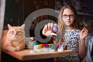 Teen girl paints Easter eggs, her ginger cat lies on the table