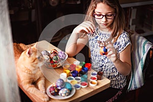 Teen girl paints Easter eggs, her ginger cat lies on the table.