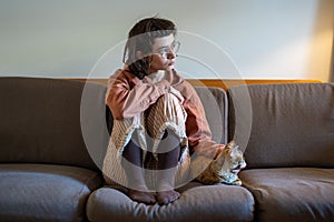 Teen girl petting cat sitting on couch at home in pyjamas looking at window at home. Pet therapy