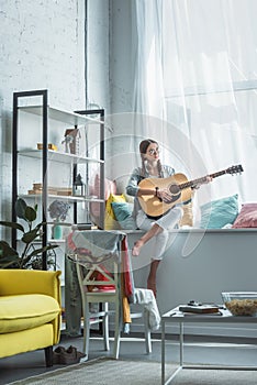 teen girl playing acoustic guitar while sitting on windowsill photo