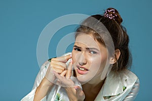 Teen girl with problem skin look at pimple, blue background. Morning theme, skincare. Teenager with acne