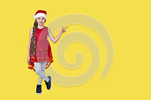 Teen girl in red dress and Santa Claus red hat standing on yellow background showing direction. Girl in Santa hat