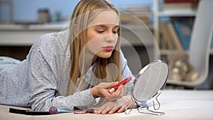 Teen girl with red glossy lipstick on lips looking in mirror, applying make up