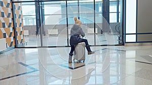 Teen girl is riding on suitcase in airport terminal waiting her flight near the escalator.