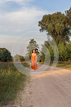 A girl in a rural summer long dress walks barefoot along a country sandy road at sunset in the village