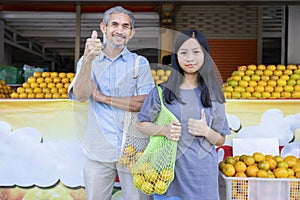 teen girl and senior grandfather carry net tole bag of oranges and doing thumbs up in front of community fruits store