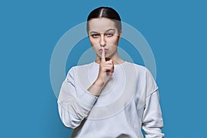 Teen girl showing silent gesture with finger on lips, blue background