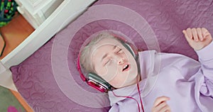 Teen girl is singing with closed eyes lying on her bed in bedroom wearing headphones