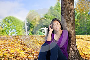 Teen girl sitting against autumn tree using cell phone