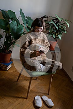 Teen girl sitting on chair with cup of tea, reading news, scrolling photos, messaging in smartphone