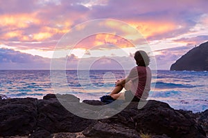 Teen girl sitting on rocky beach watching the hawaiian sunrise