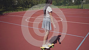 Teen girl in sneakers, skirt and t-shirt riding yellow skateboard on an outdoor basketball court. Shadow follows the