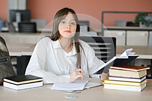 Teen girl studying with textbook writing essay learning in classroom.