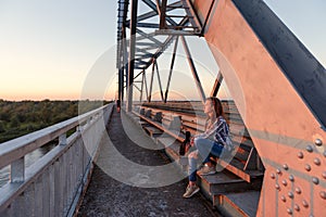 Teen girl wanderer sitting on steel bridge design in docks and looking away at sunset
