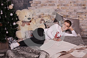 Teen guy lies on a cozy bed near a Christmas tree with a big bear