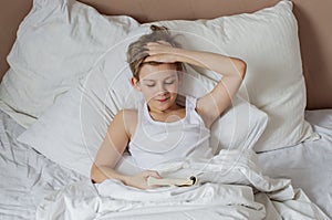 Teen handsome blonde boy lying in white bed  and reading book. Relax time