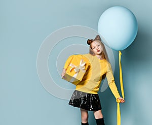 Teen kid in yellow sweatshirt, black skirt, knee-highs, boots. Kissing you, holding balloon, gift box, posing on blue background.