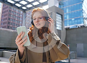 Teen redhead girl wearing headphones using smartphone in big city.