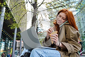 Teen redhead hipster girl using laptop talking on phone on city urban street.