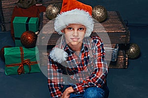 Teen in Santa hat wearing a checkered shirt sitting in a room with Christmas gifts, balls and wooden boxes.