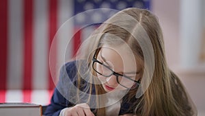 Teen schoolgirl adjusting eyeglasses reading book sitting at desk in classroom. Portrait of absorbed interested smart