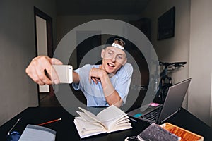 A smiling teen student doing selfie while studying at home behind a desk in the room. Selfies fees.