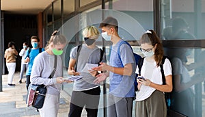 Teen students in medical masks standing with workbooks in schoolyard