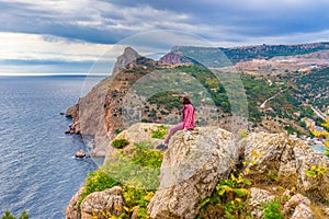 Teen tourist girl sitting on mountain cliff against beautiful seascape in cloudy day. Young lady relaxing on background of sea,